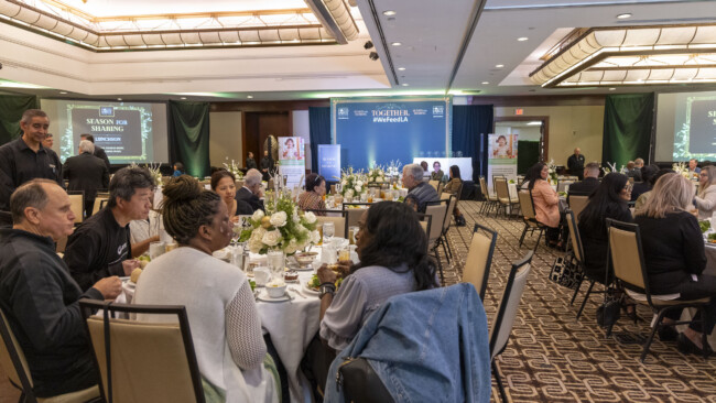 A large indoor event with round tables filled with people dining supports the Los Angeles Regional Food Bank. The stage at the front displays a "Together" banner. Warm lighting and floral centerpieces adorn each table as attendees engage in lively conversation, united for a cause.