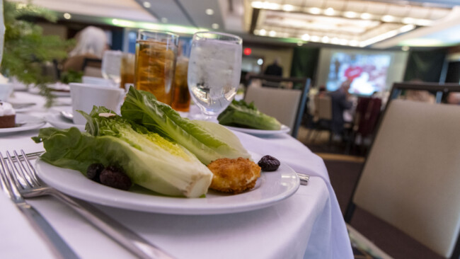 The elegant banquet table setting, provided by Los Angeles Regional Food Bank, showcases a plate with romaine lettuce, a small fried item, and black olives. Nearby are glasses of iced tea and water. In the background, a conference room is equipped with tables and a presentation screen.