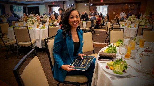 At a formal event supporting the Los Angeles Regional Food Bank, a woman in a blue blazer smiles while seated at a table. She holds a book, surrounded by plates of salad and glasses of iced tea. Other guests enjoy the gathering at elegantly set tables in the background.