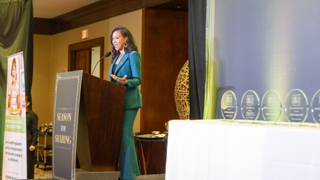 A woman in a teal suit speaks at a podium that reads "Season for Sharing," with award trophies displayed on a table to her right, highlighting the efforts of the Los Angeles Regional Food Bank.