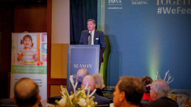 A man in a suit speaks at a podium labeled "Season of Sharing" at a formal event for the Los Angeles Regional Food Bank. Nearby, a sign depicts a child holding a bowl of food, while attendees are seated in the foreground, highlighting the community's commitment to ending hunger.