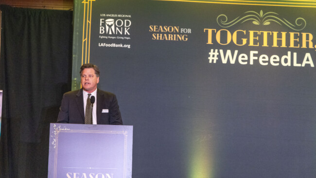 A man in a suit stands at a podium, passionately addressing an event for the Los Angeles Regional Food Bank, with "Los Angeles Regional Food Bank" prominently displayed and hashtags like "#WeFeedLA" and "Season for Sharing." The formal setting features elegant decorative elements that enhance the occasion.