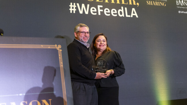 A man and a woman stand together, beaming as they hold an award, in front of a "#WeFeedLA" backdrop. Dressed in formal attire, they celebrate at an event supporting the Los Angeles Regional Food Bank.