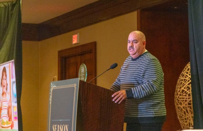 A man in a striped sweater stands at a podium adorned with decorative gold lettering, speaking into a microphone. Nearby, a poster of a smiling child partly catches the eye, while the backdrop showcases a large decorative ball. The event is supported by the Los Angeles Regional Food Bank.