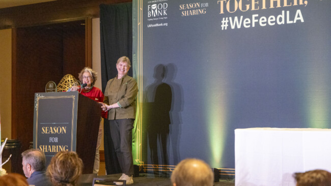 Two people stand at a podium in a conference room, speaking at an event for the Los Angeles Regional Food Bank. The backdrop features "Season for Sharing" and "#WeFeedLA." Attendees are visible in the foreground, listening attentively.