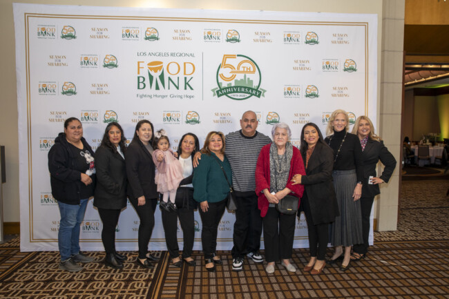 A group of people stands together, smiling in front of a Los Angeles Regional Food Bank backdrop, which prominently features their logo and a 50th-anniversary emblem. The setting is formal, highlighting the organization's commitment to fighting hunger in the community.