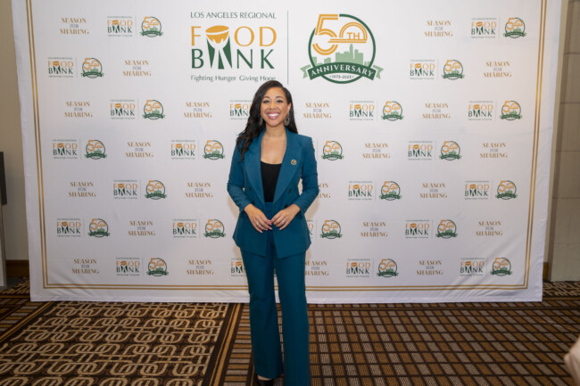 A woman in a blue suit stands smiling on a carpeted floor, framed by the Los Angeles Regional Food Bank logo and a 50th-anniversary emblem.