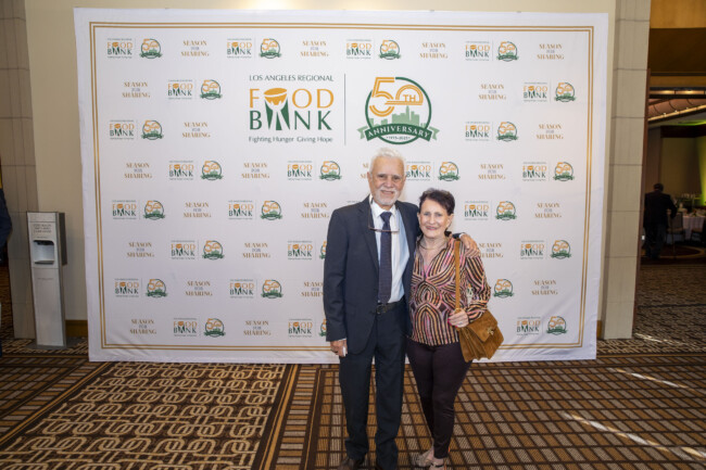 An older man in a suit and an older woman in casual attire stand smiling together in front of a backdrop featuring the Los Angeles Regional Food Bank 50th Anniversary logos, capturing the joyful spirit of this meaningful event.