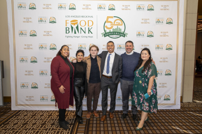 A group of six people stand together smiling in front of a backdrop featuring the "Los Angeles Regional Food Bank" and "50th Anniversary" logos. They are dressed in formal attire, and the carpet displays a geometric pattern.