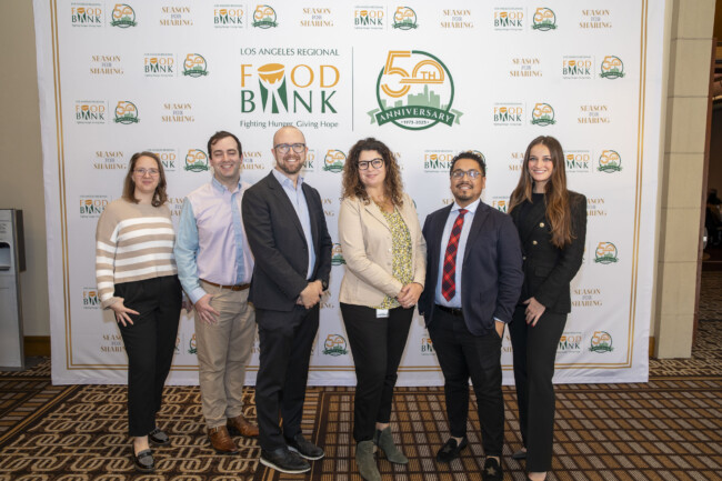A group of six people, dressed in business attire, is smiling for the camera in front of a backdrop adorned with logos celebrating the Los Angeles Regional Food Bank's 50th anniversary.