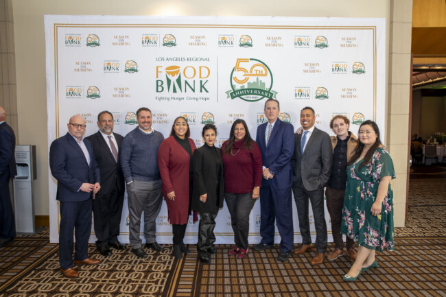 A group of people stands in front of a backdrop with the Los Angeles Regional Food Bank logo, joyfully celebrating its 50th anniversary. Dressed in both business and casual attire, they smile warmly at the camera, embodying a shared commitment to community support.