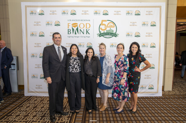 A group of six stands smiling in front of a Los Angeles Regional Food Bank's 50th anniversary banner. Dressed in business and casual attire, they pose on a patterned carpet, celebrating the impact and legacy of the food bank in their community.