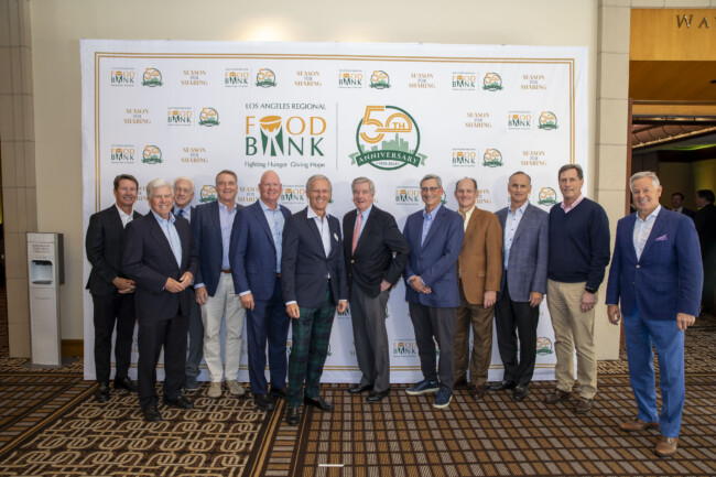 A group of people stands proudly, dressed in formal attire, on a patterned carpet. They pose in front of a backdrop celebrating the Los Angeles Regional Food Bank's 50th anniversary.