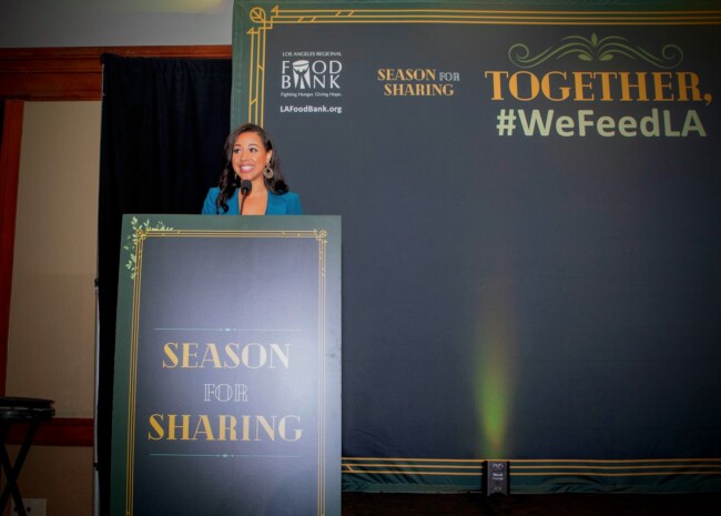 A woman stands at a podium labeled "Season for Sharing" with the Los Angeles Food Bank logo, part of the Los Angeles Regional Food Bank's initiative. She addresses an audience against a backdrop reading "TOGETHER, #WeFeedLA," highlighting community efforts to fight hunger.