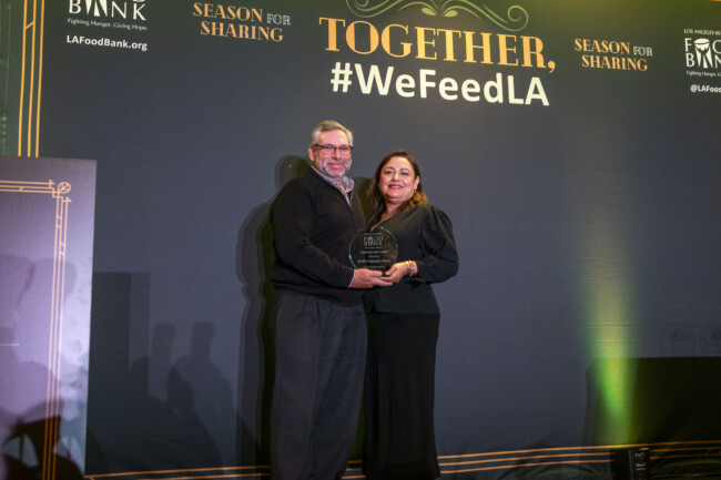 A man and a woman stand together holding an award in front of a backdrop reading "Together, #WeFeedLA" and "Season for Sharing" at the Los Angeles Regional Food Bank event. They are smiling at the camera.