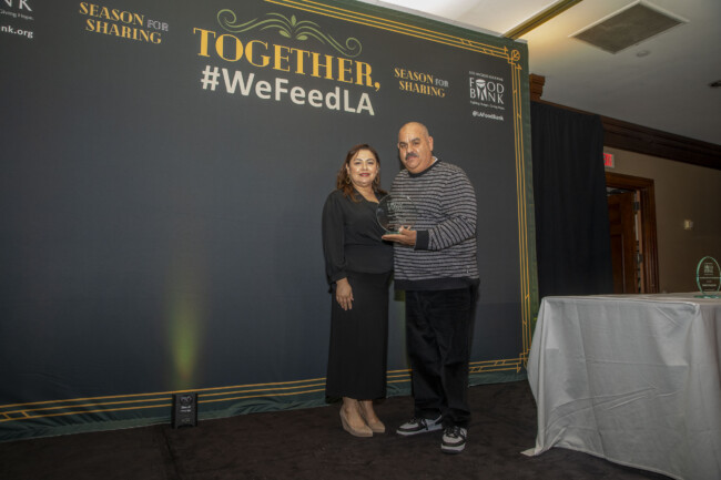 A man and woman stand on a stage in front of a backdrop with the text "Together, #WeFeedLA" and "Season for Sharing" at a Los Angeles Regional Food Bank event. The man holds an award proudly.