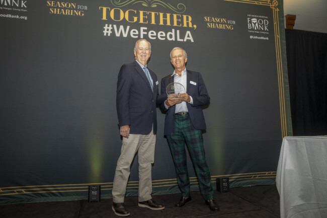 Two men stand smiling in front of a "#WeFeedLA" backdrop, proudly holding an award. Clad in suits, with one sporting plaid pants, they celebrate the success of the "Season for Sharing" initiative by the Los Angeles Regional Food Bank.