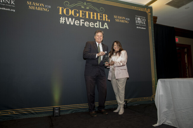 Two people stand on stage, proudly holding a glass award in front of a backdrop that reads "TOGETHER, Season for Sharing, #WeFeedLA," celebrating the efforts of the Los Angeles Regional Food Bank. They are dressed in formal attire.