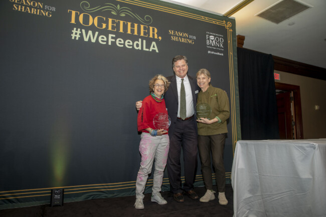 Three people stand on stage holding awards in front of a backdrop with "Together, #WeFeedLA." They smile at the camera, dressed in formal and casual attire. The event is organized by the Los Angeles Regional Food Bank.