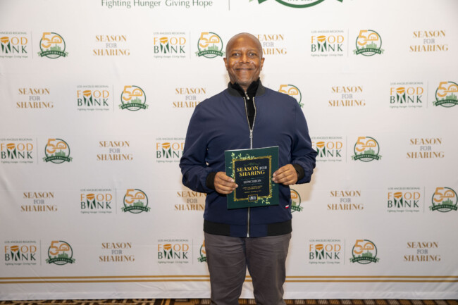 A person stands smiling, holding a "Season for Sharing" booklet in front of a backdrop adorned with "Food Bank" and "Season for Sharing" logos. They are wearing a dark jacket and grey pants, representing the spirit of giving at the Los Angeles Regional Food Bank.