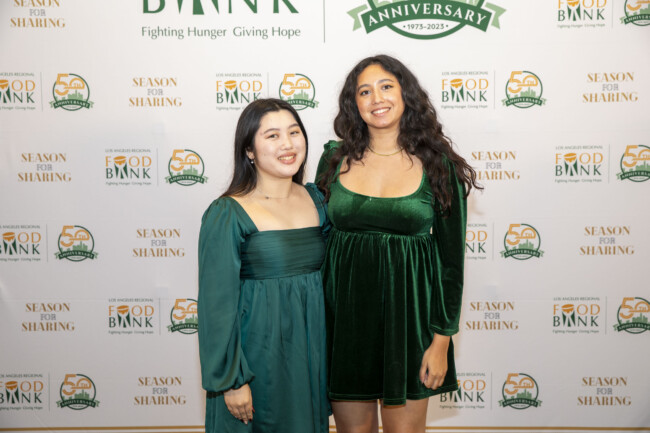 Two women in green dresses stand smiling in front of a backdrop featuring logos for the Los Angeles Regional Food Bank, "Season for Sharing," and an anniversary message. The backdrop is adorned with vibrant green and orange accents.
