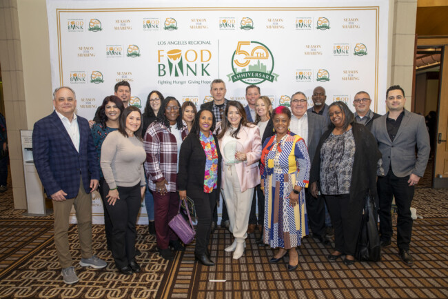 A diverse group of people stands together, beaming in front of a backdrop adorned with the Los Angeles Regional Food Bank's logo and 50th Anniversary design. Dressed in both business and casual attire, their smiles reflect a shared commitment to fighting hunger in the community.