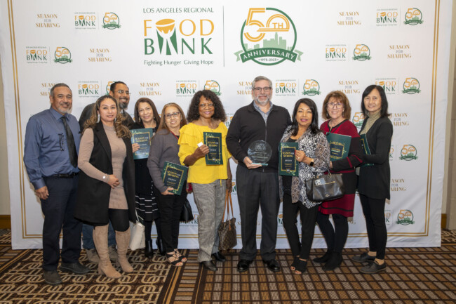 A group of people stands together, smiling, while holding plaques and an award in front of a backdrop celebrating the 50th anniversary of the Los Angeles Regional Food Bank.