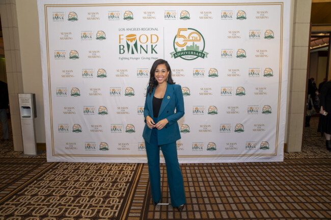 A woman in a turquoise suit poses gracefully before the Los Angeles Regional Food Bank's 50th Anniversary backdrop, adorned with its emblematic logos and celebratory text.