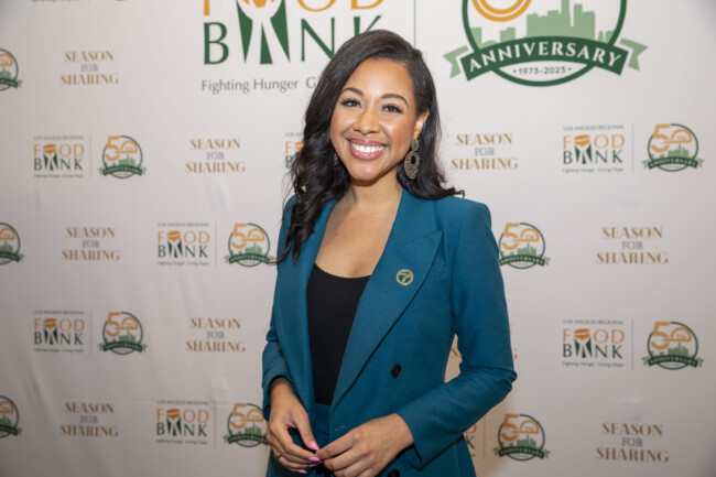 A person in a teal blazer stands smiling in front of a backdrop adorned with the Los Angeles Regional Food Bank logos, celebrating the 50th anniversary. The display features phrases like "Season for Sharing" and highlights efforts to fight hunger.