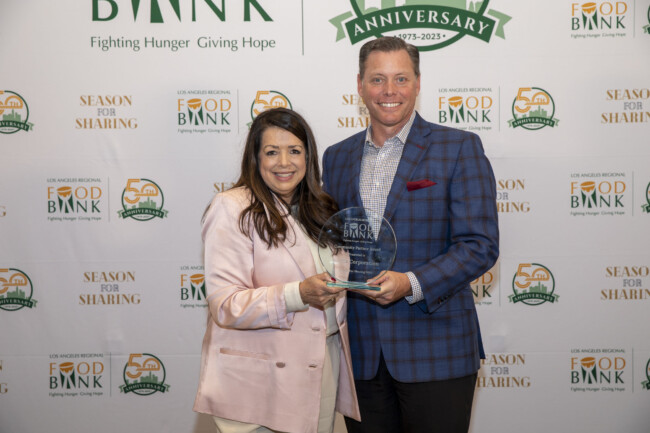 Two people stand together smiling, proudly holding an award at the Los Angeles Regional Food Bank event. The backdrop showcases logos and text celebrating the Los Angeles Regional Food Bank's anniversary and mission.
