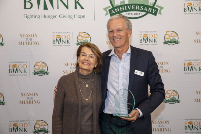 A man and a woman stand smiling together in front of a backdrop celebrating the Los Angeles Regional Food Bank's anniversary. The man proudly holds a glass award, showcasing their commitment to fighting hunger.