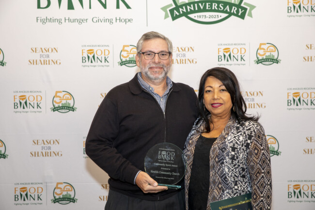 A man and woman stand together holding an award at a Food Bank event. The backdrop showcases the Los Angeles Regional Food Bank logo alongside the 50th-anniversary celebration and the text "Season for Sharing.