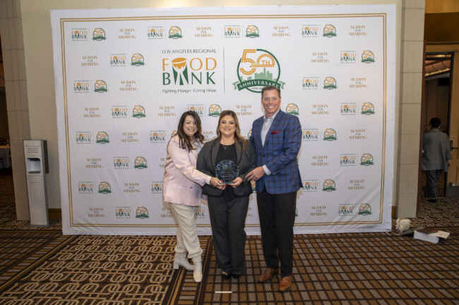 Three people stand together smiling, proudly holding an award in front of a Los Angeles Regional Food Bank 50th Anniversary backdrop. The woman in the center wears black, the person on the left is in pink, and the person on the right sports a blue plaid jacket.
