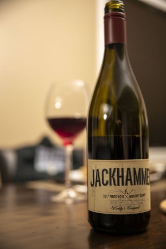 A bottle of Jackhammer 2017 Pinot Noir from Monterey County graces the wooden table, with a filled wine glass subtly suggesting indulgence. In the blurred indoor setting, it quietly echoes gatherings supporting causes like the Los Angeles Regional Food Bank.