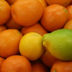 A pile of vibrant orange tangerines, sourced from the Los Angeles Regional Food Bank, with a single lemon and lime prominently displayed on top, adding a splash of yellow and green to the image.