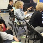 A seated woman with curly hair speaks into a microphone during a meeting at the Los Angeles Regional Food Bank. Other attendees are in folding chairs, some taking notes. The indoor setting is filled with casually dressed individuals listening attentively.