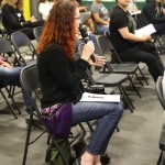 A woman with red hair, wearing jeans and a black sweater, sits on a foldable chair holding a microphone, engaged in conversation. Several people are seated around her in a casual indoor setting at the Los Angeles Regional Food Bank.