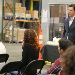 A group of people sits in chairs facing a speaker holding a microphone in an indoor setting. Nearby, a man in a suit stands by. In the background, shelves with boxes from the Los Angeles Regional Food Bank accompany a table with refreshments, underscoring their mission to combat hunger.