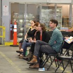 Folks seated on folding chairs in a warehouse-like setting, attentively watching something out of frame. The chairs are neatly arranged in rows against a backdrop of stocked goods on shelves at the Los Angeles Regional Food Bank, creating an informal and casual atmosphere.