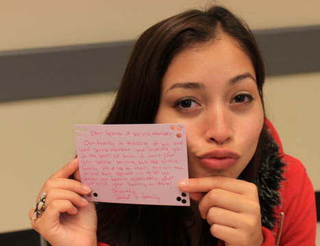 Writing letters filled with gratitude and appreciation to the families of active service members. A young woman in red holds up a written index card addressed to families of active service expressing gratitude.