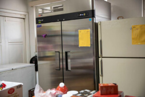 A kitchen scene features a stainless steel double-door commercial refrigerator next to a white fridge, reminiscent of setups at the Los Angeles Regional Food Bank. The area includes a countertop with assorted bottles and boxes, while a yellow paper is taped to the front of each refrigerator.