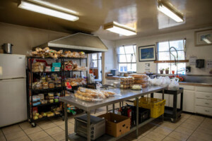 A kitchen at the Los Angeles Regional Food Bank features a metal table stacked with bread rolls and plastic bags. Shelves brimming with various baked goods line the left wall. Bright overhead lights illuminate the room, which is outfitted with white cabinets and large windows.