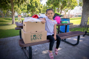 A young girl sits on a picnic table beside a large cardboard box from the Los Angeles Regional Food Bank, filled with clothes. Trees and a park setting unfold in the background, creating a serene scene of community support.