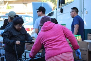 At a Los Angeles Regional Food Bank distribution event, people gather with purpose. A woman in a pink coat pushes her cart as others assist with boxes and supplies. Nearby, a truck stands ready while conversations buzz among the engaged individuals.