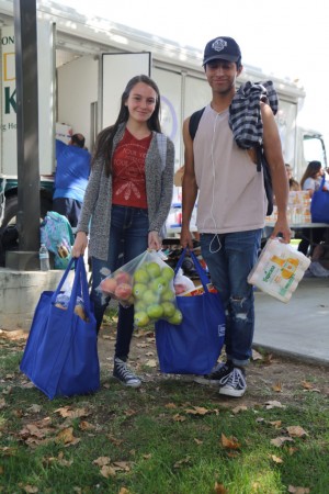 Two people stand outdoors holding reusable bags filled with groceries, including apples, smiling as a Los Angeles Regional Food Bank food truck is visible in the background. A few autumn leaves are scattered on the grass around them.