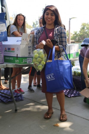 A woman smiles while holding a bag of apples and a blue tote bag at the Los Angeles Regional Food Bank. Behind her, people are filling boxes with produce at an outdoor table. There are boxes labeled "Awesome Organics" and "Arctic Apples.