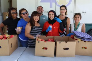 A diverse group of seven people stands behind a Los Angeles Regional Food Bank table filled with boxes of apples. They are smiling at the camera, with one person wearing a hijab and holding a can. Some are wearing sunglasses, as they proudly showcase their contribution to the community.