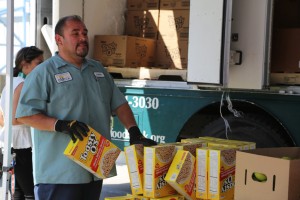 A man in a uniform and gloves is unloading boxes of "Toasty O's" cereal from a truck at the Los Angeles Regional Food Bank. Another person stands nearby amidst more boxes. The truck's open door reveals shelves stocked with supplies ready to support those in need.
