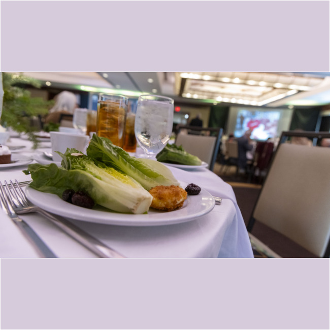 The elegant banquet table setting, provided by Los Angeles Regional Food Bank, showcases a plate with romaine lettuce, a small fried item, and black olives. Nearby are glasses of iced tea and water. In the background, a conference room is equipped with tables and a presentation screen.