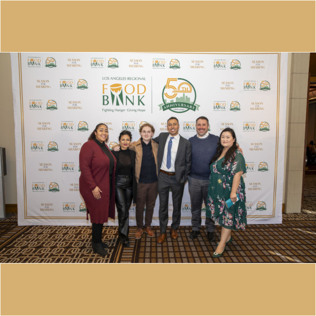 A group of six people stand together smiling in front of a backdrop featuring the "Los Angeles Regional Food Bank" and "50th Anniversary" logos. They are dressed in formal attire, and the carpet displays a geometric pattern.